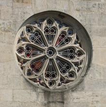Teilansicht einer Kirchenfassade mit achtflügeliger Fensterrosette aus hellbraunem bis hellgrauem Gestein.