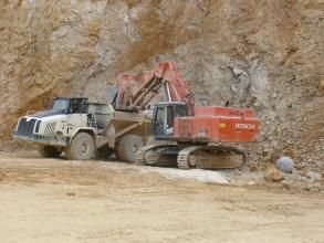 Das Bild zeigt einen orangenen Bagger und ein Transportfahrzeug vor einer Abbauwand in einem Steinbruch. Der Bagger lädt gerade Material auf das Fahrzeug.
