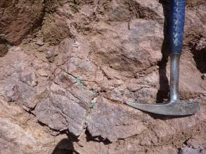 Teilansicht von rötlich grauem bis braunem Gestein, teils als Bruchschollen ausgebildet. In einer Gesteinsfuge unterhalb der Bildmitte ist eine grünliche Verfärbung erkennbar. Rechts ist ein Geologiehammer angelehnt.