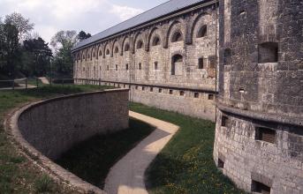 Blick auf eine große Festungsanlage aus grauem Mauerwerk, teils schwärzlich verfärbt, mit Wallmauer und Graben, langem Hauptbau und rechts noch angeschnittenem Rundbau.