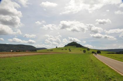 Der Zeugenberg Hohenkarpfen bei Hausen ob Verena (Lkr. Tuttlingen) Das Bild zeigt weite, braun und grün gefärbte Ackerflächen, an die sich eine kegelförmige, oben bewaldete Erhebung anschließt. Links und rechts sind weitere, jedoch flachere bewaldete Höhenzüge erkennbar. Am Himmel ziehen lockere Wolken vorbei.