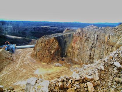 Spaltenfüllung in der Hauptrogenstein-Formation, Steinbruch der Fa. Hauri, Merdingen Blick von oben über eine Gesteinskante in einen Steinbruch. Zu sehen ist eine rundliche, nach rechts abknickende rötlich braune Steinbruchwand. Der Knick zur rechts steileren und höheren Wand ist mit blaugrauem Material verfüllt.