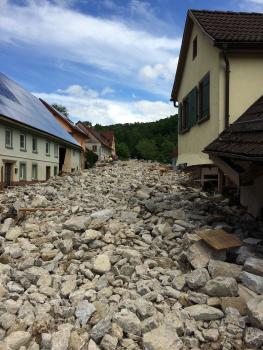 Ablagerungen der Geröllflut in der Ortslage von Braunsbach (Foto: LRA Schwäbisch Hall, 06/2016) Blick von vorne auf eine Straße einer Ortschaft, welche vollständig mit Geröll gefüllt ist.