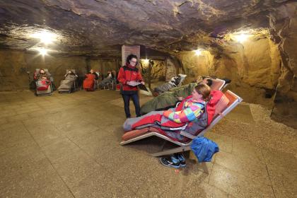 Besucherbergwerk Tiefer Stollen – Ein Blick in die Therapiehalle unter Tage; Foto: Stadt Aalen/Besucherbergwerk Tiefer Stollen Blick in die Halle eines ehemaligen Bergwerkes. In dem gut ausgeleuchteten, mit Bodenfliesen bedeckten Raum sind mehrere Liegstühle aufgestellt. Die Stühle sind mit Menschen in Warmhaltedecken besetzt.