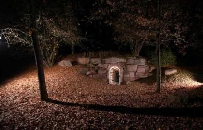 Eingangsbereich beim Silberbergwerk Himmlisch Heer; Foto: Bergwerk Hallwangen In einer Bodensenke, von Steinquadern und Bäumen umgeben, liegt die schmale Eingangstür eines Bergwerks. Das Foto entstand bei Dunkelheit, die vergitterte Eingangstür ist jedoch beleuchtet.