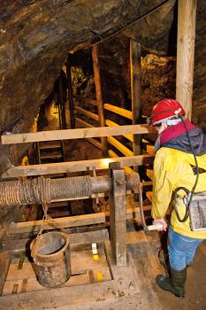 Besucherbergwerk Grube Wenzel bei Oberwolfach; Foto: Gemeinde Oberwolfach Blick in das Innere eines Bergwerks. Gezeigt wird hier ein Förderkorb mit Winde und Handkurbel, die eine Person in Schutzkleidung bedient. Im Hintergrund ein abgestützter Stollen sowie ein Aufstiegsschacht.
