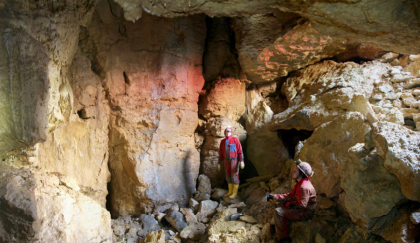 Das Bild zeigt zwei Personen in Schutzanzügen und mit Helm, die in einer Höhle stehen. Sie sind umgeben von hellbraunem bis gelblichem Gestein. 