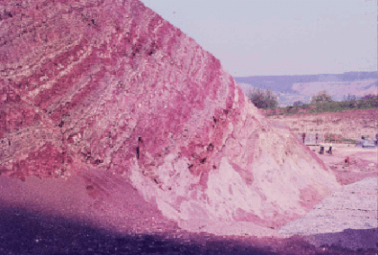 Antithetisch rotierte Rutschscholle in der Einschnittsböschung am Wildenberger Hang (Foto: Brodbeck, 2010) Das Bild zeigt zentral und rechts eine aufgeschlossene Gesteinsformation, die eine rötliche Farbe hat. Sie ist geschichtet mit einzelnen weißen Bändern. Dahinter befinden sich plateauartige Berge.