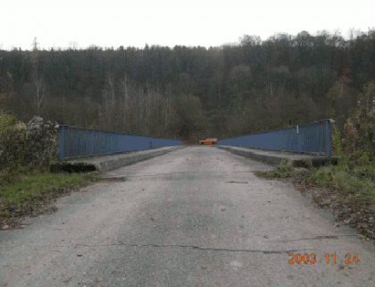Risse im Fahrbahnbelag einer Feldwegüberführung (Foto: Brodbeck, 2010) Zu sehen ist eine Straße, die geradewegs auf eine Brücke führt. Hinter der Brücke befindet sich ein bewaldeter Hang.