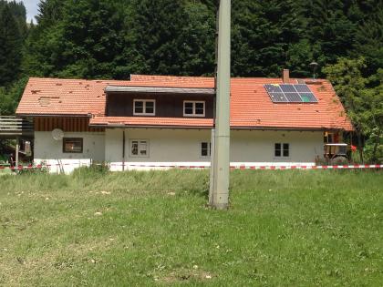 Schäden an benachbarter Bebauung (Foto: WWA Kempten, 2015) Der Blick richtet sich von einer Wiese auf ein Haus. Das Haus weist einige Beschädigungen, so wie fehlende Dachziegel oder Einschläge in der Häuserwand auf.