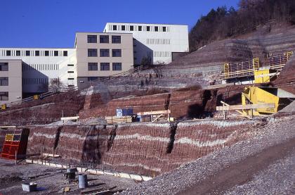 Mittlerer Keuper, Tübingen Mehrstufige, nach rechts hin ansteigende Baugrube vor modernen Gebäuden aus in Schichten gelagertem, dunkelrotem und weißem Gestein.