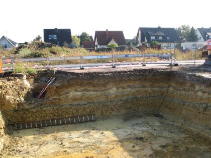 Pechelbronn-Formation (Oberrheingraben-Tertiär), Gottenheim Rechteckige Braugrube, in der helles, geschichtetes Sedimentgestein ansteht. Dahinter befinden sich einige Wohnhäuser.