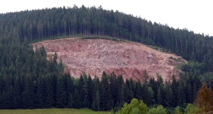 Granitsteinbruch im Eisenbach-Granit bei Hammereisenbach Weiter Blick auf einen langgezogenen, oben und unten dicht bewaldeten Berghang mit in der Mitte offener Flanke. Das hier in einem Steinbruch aufgegrabene und abgetragene Gestein ist rötlich grau.