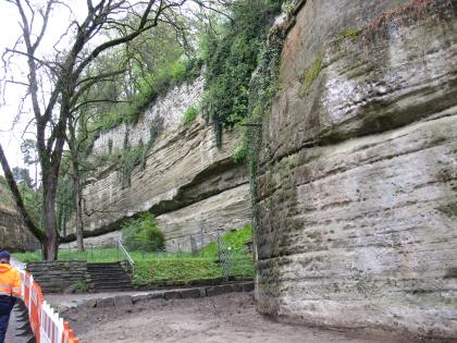 Auf dem Bild sieht man auf der rechten Seite eine Felswand aus geschichtetem Sedimentgestein. Auf der linken Seite vor dem Aufschluss befinden sich zwei Bäume, sowie ein Weg. Der Weg ist durch eine Absperrung versperrt.