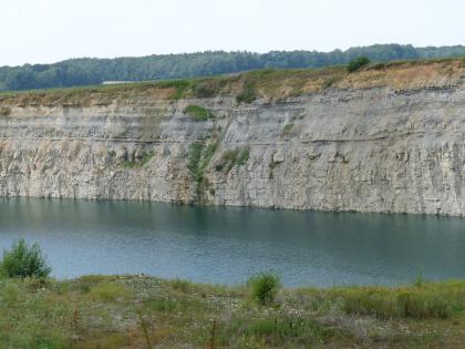 Aufgelassener Steinbruch Schmalfelden Hinter einer begrünten Kuppe im Vordergrund und einem blauen See darunter steigt die waagrecht gebankte, grauweiße Wand eines Steinbruchs auf. Das obere Ende des Bruchs ist braun gefärbt und mit Gras bewachsen.