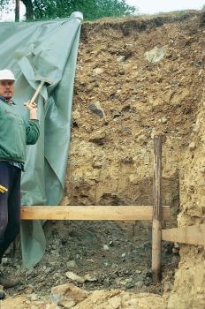 Baugrube an einem südost-exponierten Unterhang im Neckartal bei Horb-Dettingen Das Bild zeigt die über 2 m hohe Wand einer Baugrube. Die gelblich braun gefärbte Grube ist mit Steinen durchsetzt und von einer Absperrung gesichert. Am Rand der Grube schlägt ein Arbeiter eine Plane zurück.