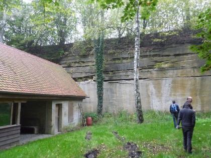 Lettenkeuper-Hauptsandstein in Flutfazies Hinter einem niedrigen Gebäude links und zwei schlanken Bäumen in der Mitte ragt die Wand eines aufgelassenen Steinbruchs empor. Während dabei die untere, hellgraue Hälfte glatt erscheint, durchziehen die obere, dunkelgraue Hälfte zahlreiche Rillen.