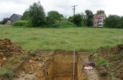 Pseudovergleyter Pelosol aus tonreicher Fließerde über Gesteinszersatz des Unteren Muschelkalks (b128) unterhalb der Ruine Hochburg (Emmendingen) Blick auf ein offenes Bodenprofil des LGRB. Das Profil, von dem drei Wände sichtbar sind, befindet sich im Vordergrund auf einer Wiese. Links und rechts des Profils liegen Erdhaufen. Im Hintergrund sind einzelne Gebäude zu sehen.