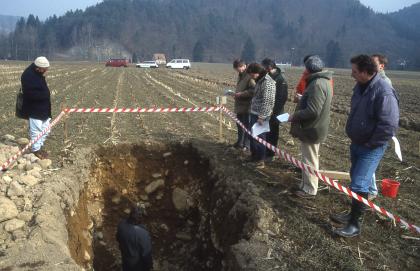 Tief entwickelte Braunerde aus schluffig-sandiger Decklage über Niederterrassenschotter Das Bild zeigt ein zum Vordergrund hin offenes Bodenprofil auf einer Ackerfläche. Das Profil ist dunkelbraun und mit Gestein durchsetzt. Links liegt Aushub. Das Profil ist mit einem farbigen Band gesichert. Mehrere Personen umstehen das Profil.