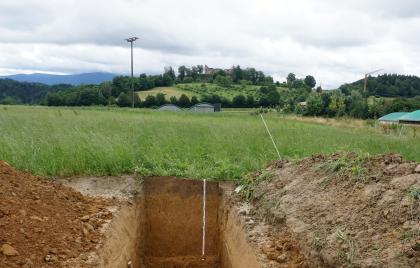 Tief entwickelte pseudovergleyte erodierte Parabraunerde aus jüngerem über älterem Lösslehm, unterhalb der Hochburg bei Emmendingen Blick auf ein aufgegrabenes Musterprofil des LGRB. Das Profil, von dem drei Wände sichtbar sind, liegt mittig im Vordergrund vor einer Wiese. Rechts und links ist Bodenaushub. Im Hintergrund, auf einer bewaldeten Anhöhe, liegt eine Burgruine.
