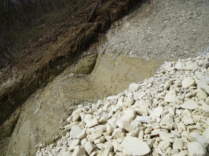 Abgetreppte Profilierung der Böschungsaufstandsfläche unterhalb der Filterschicht aus Schroppen Man schaut von der Seite auf einen steilen Hang. Im Vordergrund liegt grober Schotter auf dem Hang. Dahinter sind mehrere Stufen in den Hang eingearbeitet, der Untergrund ist verfestigt.