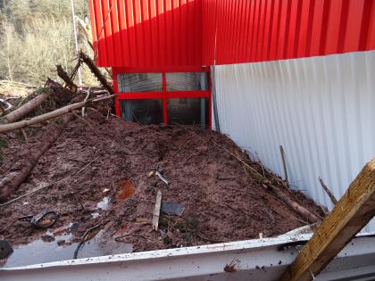 Durch Schlamm eingedrückte Gebäudewand Die rechte Seite des Bildes zeigt eine Gebäudewand aus Wellblech. Man schaut auf ein Fenster des Gebäudes. Vor der Wellblechwand befinden sich große Mengen an Schlamm mit mitgerissenem Material, die die Gebäudewand eindrücken.