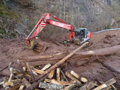 Herstellung eines Entwässerungskanals Das Bild zeigt einen roten Bagger, der versucht, mit seiner Schaufel einen Bereich vor einer Felswand vom Schlamm zu befreien. Im Vordergrund liegen ein großer Baumstamm und einige zersägte Stämme und Äste auf einem Haufen.