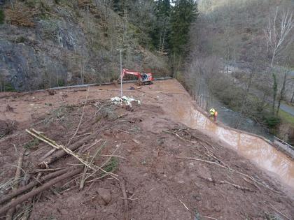 Blick auf die Murgangablagerung Aus der Vogelperspektive richtet sich der Blick auf einen Bereich, der größtenteils von großen Mengen an Schlamm verdeckt ist. Auch liegen in den Schlammmassen viele Baustämme und Äste. Ein Bagger räumt den Schlamm beiseite.