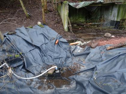 Pumpensumpf mit Tauchpumpe Zu sehen ist eine Mulde, die mit schwarzer Folie ausgelegt ist. In der Mitte der Mulde befindet sich eine Pumpe. Drumherum schauen einige Rohre aus dem Boden, aus denen Wasser in die Mulde läuft. Im Hintergrund stehen Bäume und ein Überstand aus Beton.