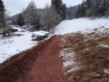Fertiggestellte Drainage Das Bild zeigt eine verschneite Landschaft. Bäume und ein Gebäude befinden sich im Hintergrund. Im Vordergrund ist rechts eine Wiese am Hang, links im Bild ein kleiner aufgeschütteter Wall. Davor befindet sich rötlicher Kies am Boden.