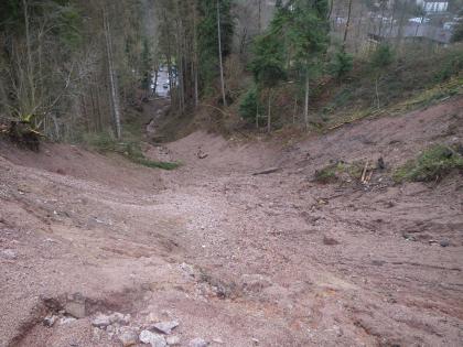 Blick in die Hangklinge Der Blick geht talabwärts vom oberen Ende eines Hanges, der mit rötlich-braunem Erdmaterial bedeckt ist und keinen Bewuchs aufweist. Weiter unterhalb säumen Bäume eine eingeschnittene Rinne, in der Wasser talabwärts fließt. Ganz unten ist ein Parkplatz.