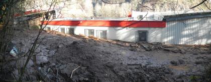 Murgangablagerung hinter dem Verbrauchermarkt Das Bild zeigt ein großes Gebäude vor dem sich einigen Mengen an Schlamm mit Trümmerteilen, Baumstämmen, Ästen und weiterem Material auftürmen.