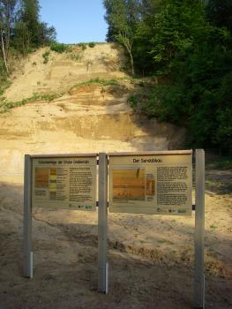 Aufgelassene Sandgrube im Gewann Grafenrain nördlich von Mauer Blick auf die hellbraune Abbauwand einer Sandgrube, die links und rechts von Wald begrenzt wird. Im Vordergrund verweisen zwei Schautafeln auf den Sandabbau und die Schichtenfolge in der Grube.