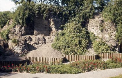 Posidonien-Schieferwand (aufgelassener Steinbruch) im Kurgebiet von Bad Schönborn Blick auf eine alte Steinbruchwand, die oben und auch zwischen den Gesteinsabschnitten bewachsen ist. Im Vordergrund verläuft ein Weg mit Blumenbeeten.