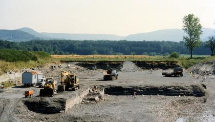 Aufgelassener Steinbruch in der Posidonienschiefer-Formation östlich von Ohmden (Lkr. Esslingen) Das Bild zeigt einen aufgelassenen Steinbruch mit nur noch niedrigen Wänden, Schutthaufen, Baufahrzeugen und Besuchern. Im Hintergrund Wald und bewaldete Höhen.