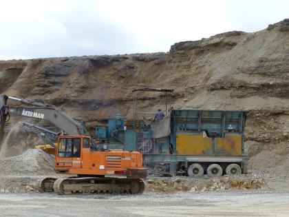 Aufbereitung des abgebauten Schotters in der Kiesgrube östlich von Zwiefaltendorf Blick auf die nach rechts ansteigende Wand einer Kiesgrube. Das Material der Wand ist bräunlich grau und zeigt waagrecht verlaufende Abstufungen. Vor der Wand steht rechts eine Fördermaschine, links ein Bagger.