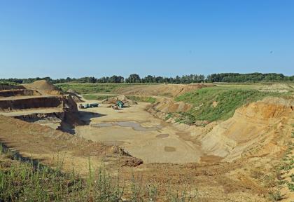 Blick über die Kiesgrube zwischen Laupheim und Burgrieden mit aktueller Abbauwand, Materialaufbereitung und Verfüllung früherer Abbaubereiche Blick über den Rand einer Kiesgrube. Links sind treppenartige Abraumflächen, rechts und im Hintergrund abgeschrägte, teils bewachsene Grubenwände zu sehen. Dazwischen breitet sich der flache Boden der Grube aus.