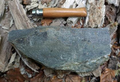 Dunkler, leicht angewitterter Amphibolit im Steinbruch auf der Südseite des Fuchsköpfles bei Freiburg-Herdern Nahaufnahme eines dunklen, bläulich grauen Gesteinsstückes. Die Kante oben verläuft fast gerade, die unten links in einem Bogen. Über dem Stück liegt ein Werkzeug als Größenvergleich.