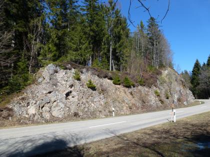 Straßenaufschluss zwischen Wolpadingen und Horbach, Dachsberg (Südschwarzwald) Zu sehen ist hier eine Straßenböschung mit offenliegendem Gestein. Die Böschung ist links der Mitte rundlich eingekerbt, außerdem fällt sie zu beiden Seiten steil ab. Die Kuppe ist bewaldet.
