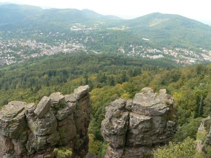 Blick vom Battertfelsen über Baden-Baden zum Fremersberg Von erhöhtem Standort aus blickt man über die abgeflachten Decksteine zweier Felstürme auf darunter sich ausbreitende Wälder. Am Fuß von bewaldeten Bergen im Hintergrund liegt verstreut eine größere Siedlung.
