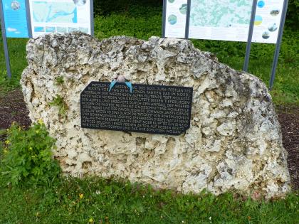 Das Bild zeigt einen großen, auf einer Wiese stehenden gelblich grauen Felsbrocken. Am Gestein ist eine schwarze Infotafel befestigt. Im Hintergrund stehen zwei größere Informationstafeln.
