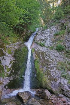 Blick auf einen fast senkrechten, oberhalb der Bildmitte nach rechts abknickenden Wasserfall. Der Wasserfall verläuft in einer schmalen Rinne zwischen hohen Felswänden. Die Felsen sind teils bemoost, links auch mit Bäumen bewachsen.