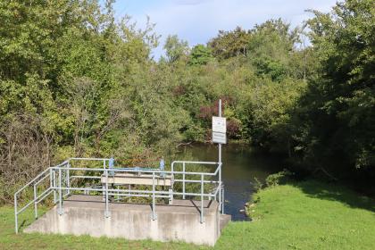 NSG Taubergießen – Einleitung von Wasser in das Gebiet außerhalb des Hochwasserdamms Blick auf eine Wehranlage mit Podest und umlaufendem Gitter. Das abgesperrte Wehr leitet Wasser in einen dahinter liegenden, von Wald umgebenen Fluss.