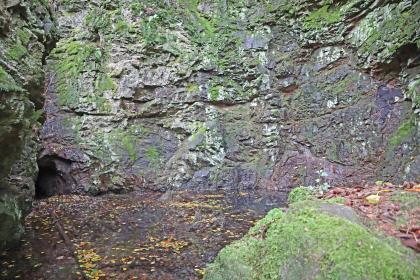 Radstube am Bergbaupfad Ehrenstetter Grund Blick auf eine mehrfarbige Gesteinswand, teils violettblau bis grau, teils grün durch Moos verfärbt. Links in der Ecke, knapp über dem Boden, ist ein kleiner Schacht erkennbar.