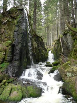 Menzenschwander Wasserfall, St. Blasien-Menzenschwand Entlang einer schmalen Felsenklamm fließt ein mehrstufiger Wasserfall, der zum Betrachter hin breiter wird. Links oben stürzt ein weiterer, sehr dünner Wasserfall von der Spitze eines Felsens herunter. Rechts führt ein Treppenweg an den Fällen entlang.