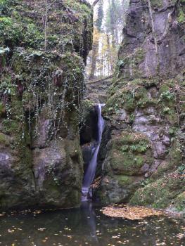 Unterer Haselbach-Wasserfall mit Teufelskessel, Gurtweil Blick auf zwei rötlich graue, stark bewachsene und bemooste Felsformationen, die am unteren Ende von einem See umschlossen sind. Etwa auf halber Höhe zwischen den Felsen stürzt sich ein schmaler Wasserfall in den See.