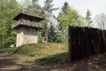 Rekonstruierter Limes-Wachturm südlich von Großerlach-Grab (Rems-Murr-Kreis) Das Bild zeigt einen am Waldrand stehenden rekonstruierten römischen Wachturm links sowie einen hölzernen Palisadenzaun rechts.