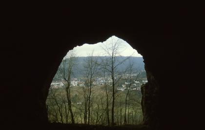 Blick aus der Rusenschlosshöhle ins Blautal bei Blaubeuren-Gerhausen Die schwarz eingerahmte Öffnung einer Höhle gibt den Blick frei auf eine Landschaft mit hoch stehenden Bäumen, tief liegenden Feldern, einer Siedlung und angrenzendem Wald.