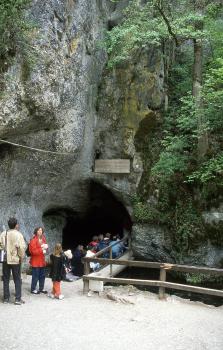 Am Eingang zur Wimsener Höhle bei Hayingen Blick auf den Eingang einer Höhle. Die Höhle liegt am unteren Ende einer hoch aufragenden Felswand. Rechts ist der Fels bewachsen. Vor dem Höhleneingang warten mehrere Menschen.