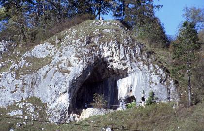 Das Bild zeigt einen hügelartig geformten, dreieckigen Felsen, dessen Kuppe mit Bäumen und Sträuchern bewachsen ist. Das bemooste, weißlich graue Gestein weist an der Vorderseite einen hohen und breiten Höhleneingang auf.